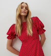 A woman with long blonde hair wears the Mela Red Polka Dot Crepe Midi Dress, featuring puffed short sleeves and an empire waist, as she stands confidently against a white wall looking at the camera.