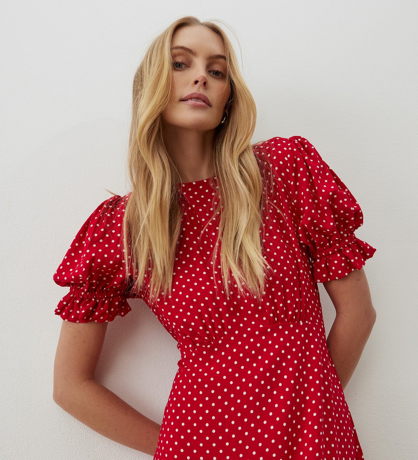 A woman with long blonde hair wears the Mela Red Polka Dot Crepe Midi Dress, featuring puffed short sleeves and an empire waist, as she stands confidently against a white wall looking at the camera.