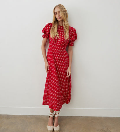 A woman stands against a white wall wearing the Mela Red Polka Dot Crepe Midi Dress, featuring an empire waist and ruffled short sleeves, styled with white ankle-tie shoes and her long blonde hair.