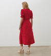 A woman with long blonde hair wears the Mela Red Polka Dot Crepe Midi Dress, featuring an empire waist, and beige wedge sandals, standing with her back to the camera in a minimal indoor setting.