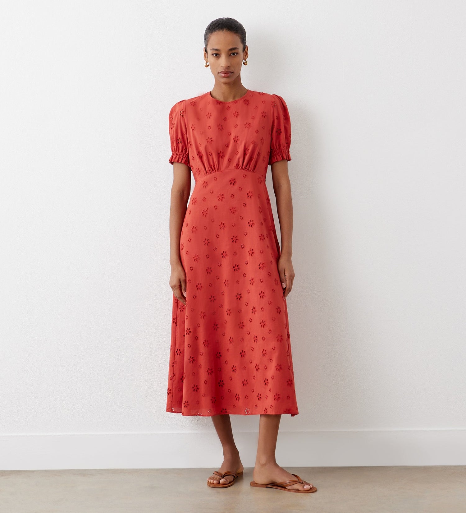 A woman stands against a white wall wearing the Mela Rust Broderie Anglaise Cotton Midi Dress with brown sandals. Her hair is pulled back, and she looks at the camera.
