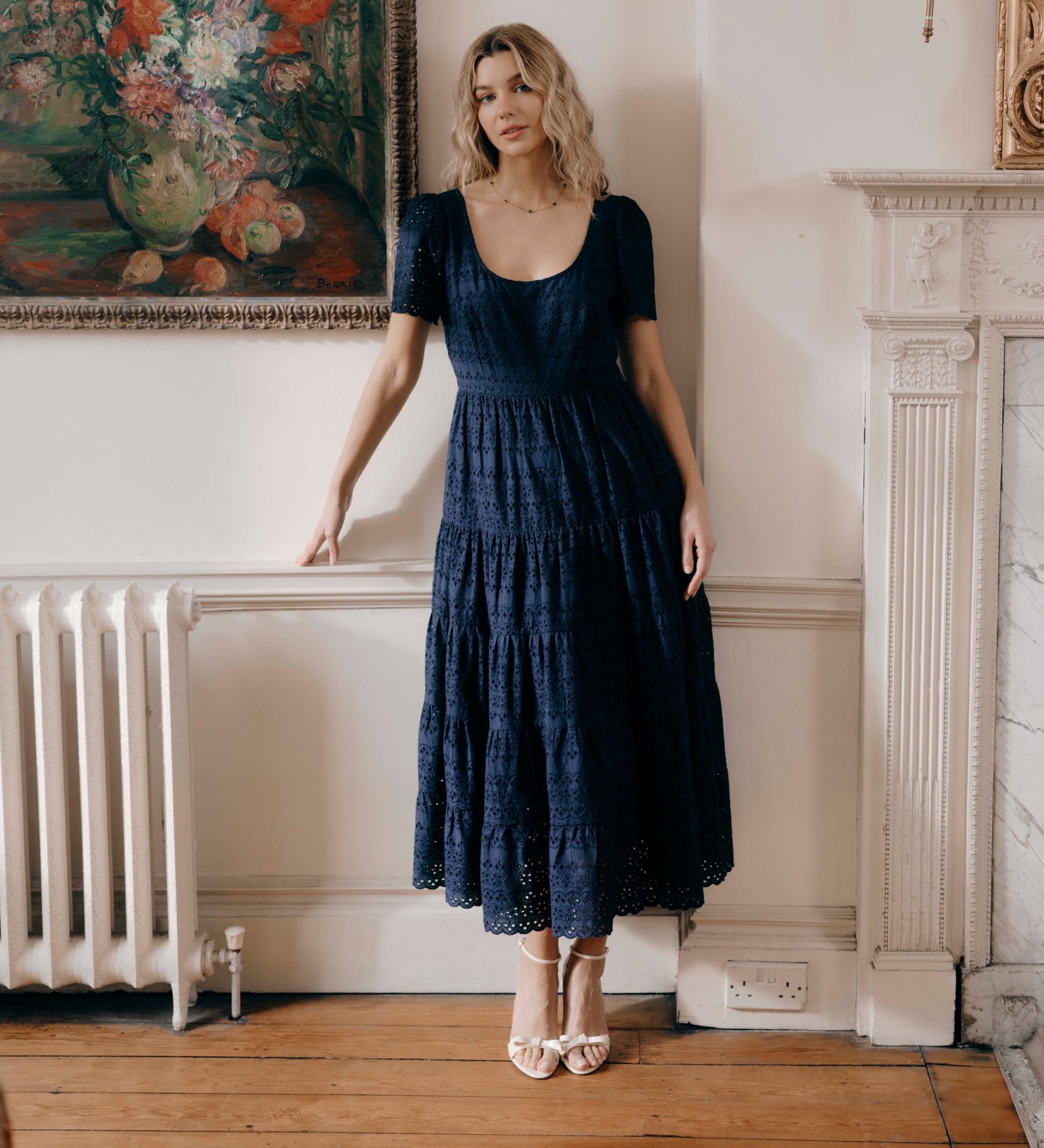 Woman in Jemma Navy Cotton Broderie Square Neck Tiered Midi Dress standing in a room with a painting and radiator in the background.