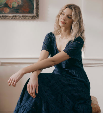 Woman in Jemma Navy Cotton Broderie Square Neck Tiered Midi Dress sitting on a couch in a room with a painting on the wall.