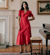 Woman in Quinn Red Cotton Broderie Lace Trim V-Neck Midi Dress standing in a room with a piano and fireplace.