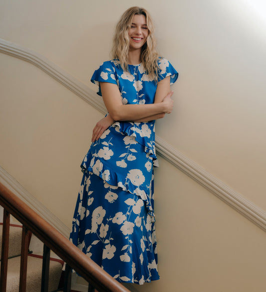 Woman in Sierra Blue Flower Asymmetric Frill Midi Tea Dress standing indoors against a beige wall.