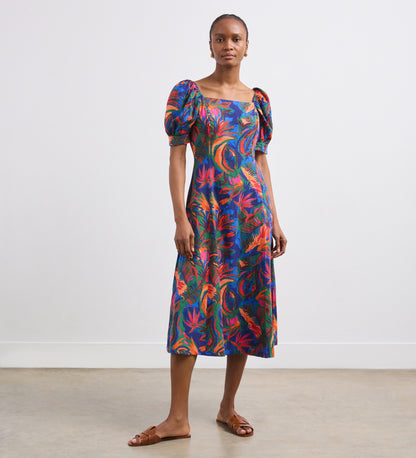 A woman stands indoors against a white wall and concrete floor, neutrally posing for the camera in the Daisy Green Tropical Linen Blend Midi Dress with puffed short sleeves and a vibrant floral print, paired with brown sandals.