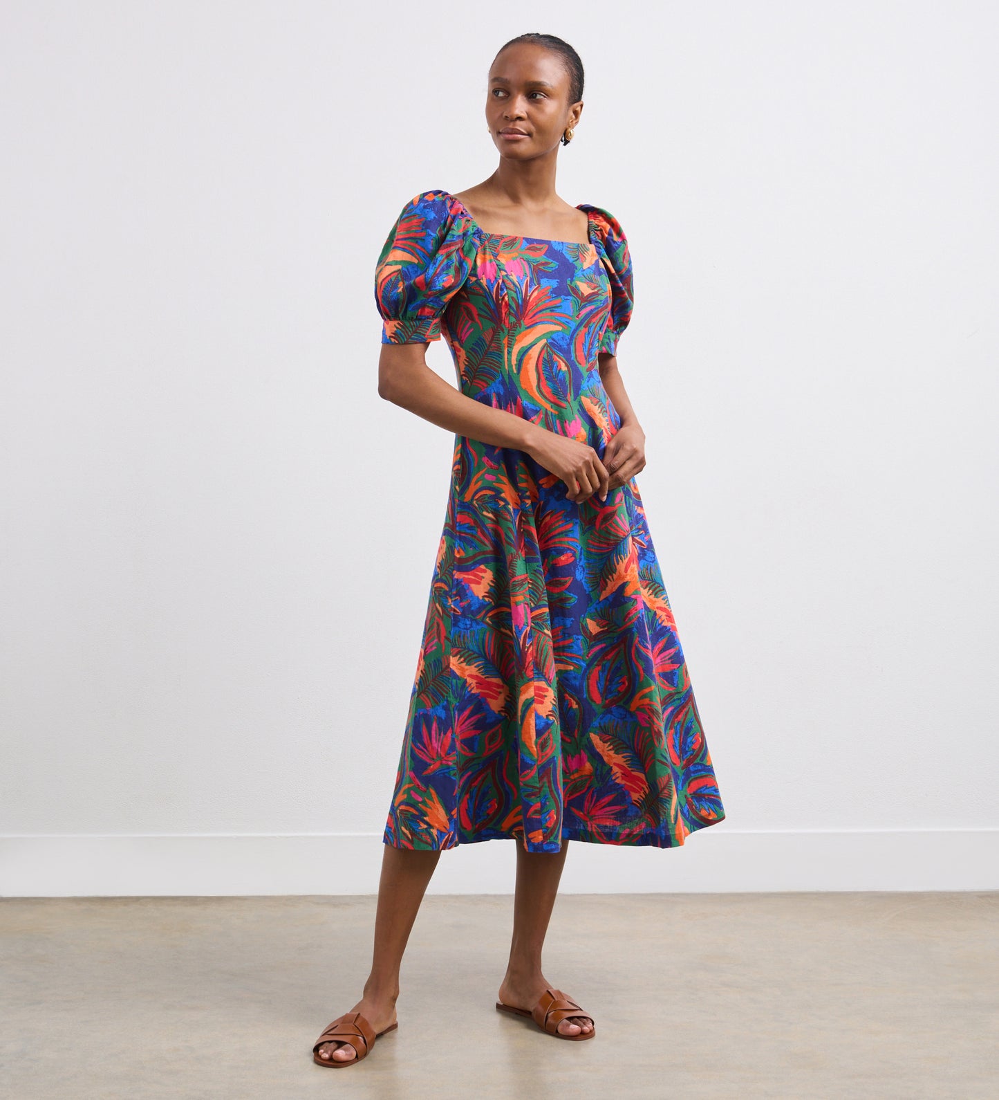 A woman stands against a white wall wearing the Daisy Green Tropical Linen Blend Midi Dress with puffed short sleeves and brown flat sandals, looking slightly to the side with her hands clasped in front of her.