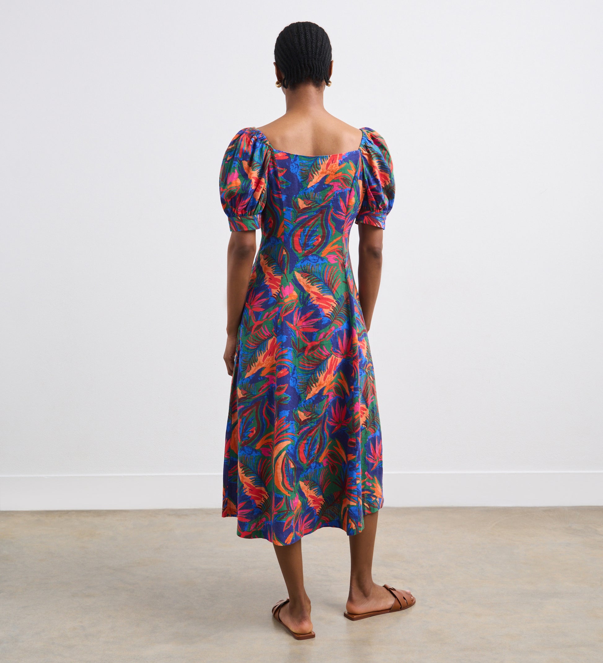 A person stands indoors facing away, wearing the Daisy Green Tropical Linen Blend Midi Dress with puffed short sleeves and flat sandals. The dress’s vibrant print stands out against the plain white walls and concrete floor.