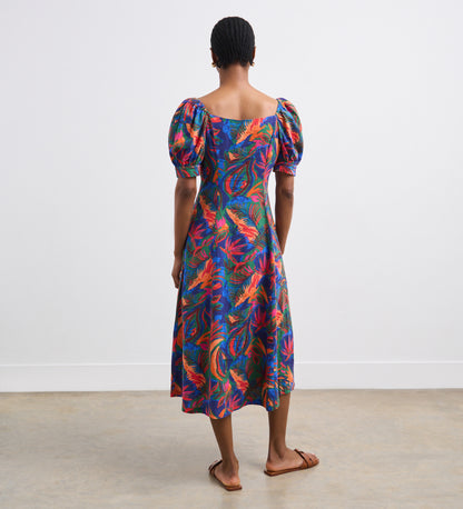 A person stands indoors facing away, wearing the Daisy Green Tropical Linen Blend Midi Dress with puffed short sleeves and flat sandals. The dress’s vibrant print stands out against the plain white walls and concrete floor.