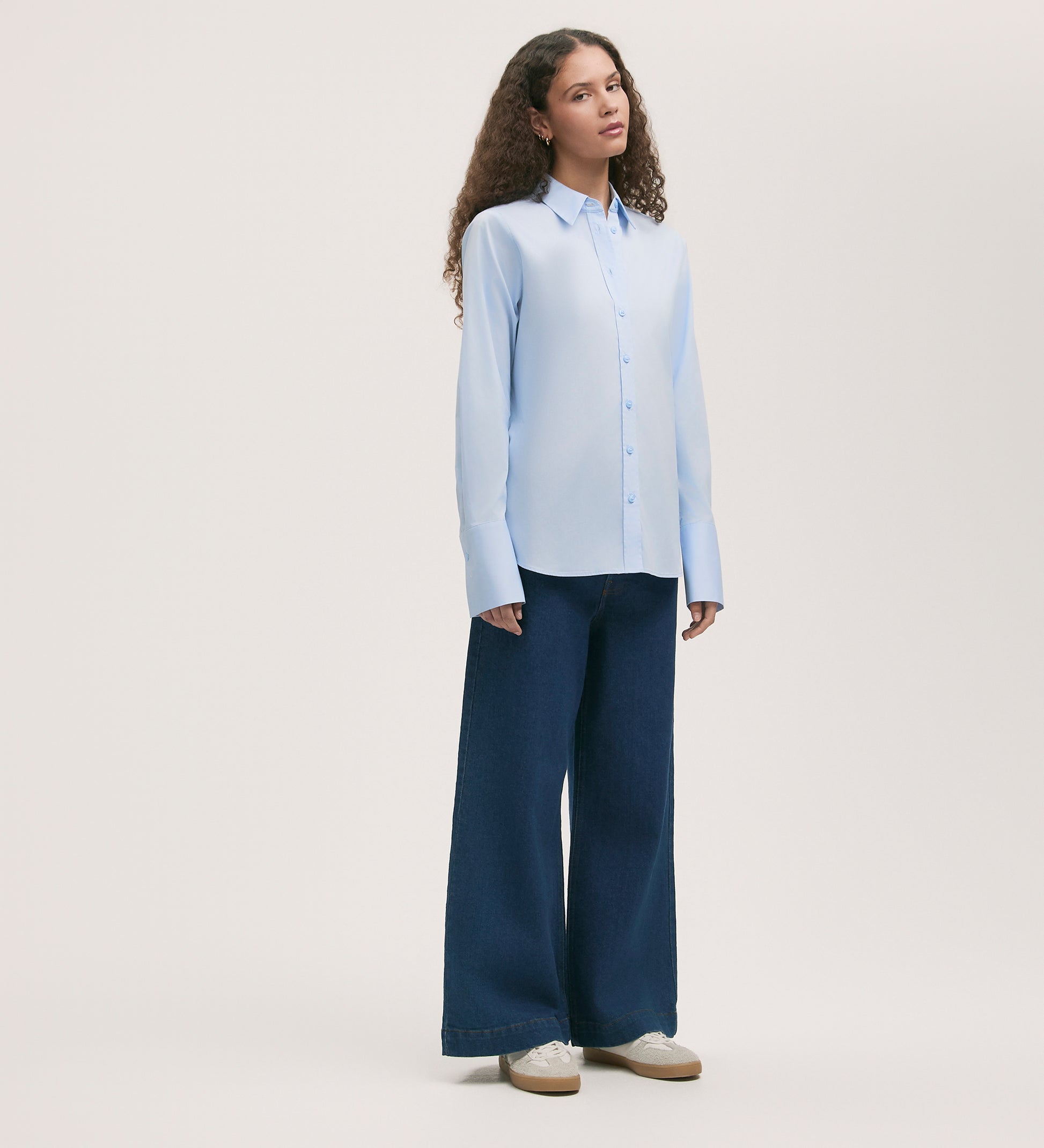 A woman with long curly hair and a neutral expression stands against a light background wearing the Dillon Light Blue Cotton Shirt, wide-leg dark blue jeans, and white sneakers.