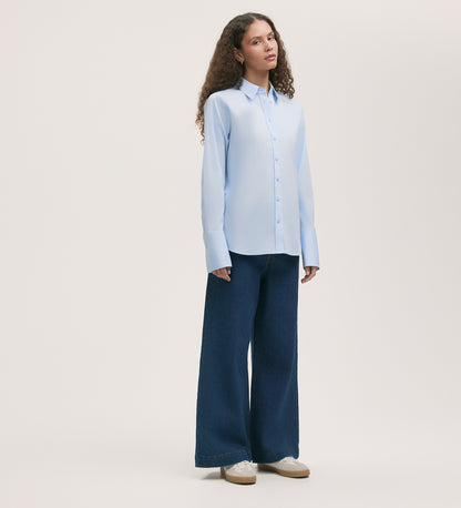 A woman with long curly hair and a neutral expression stands against a light background wearing the Dillon Light Blue Cotton Shirt, wide-leg dark blue jeans, and white sneakers.