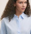A woman with curly brown hair wears the Dillon Light Blue Cotton Shirt in a regular fit, gazing slightly to the side with a neutral expression against a plain background.