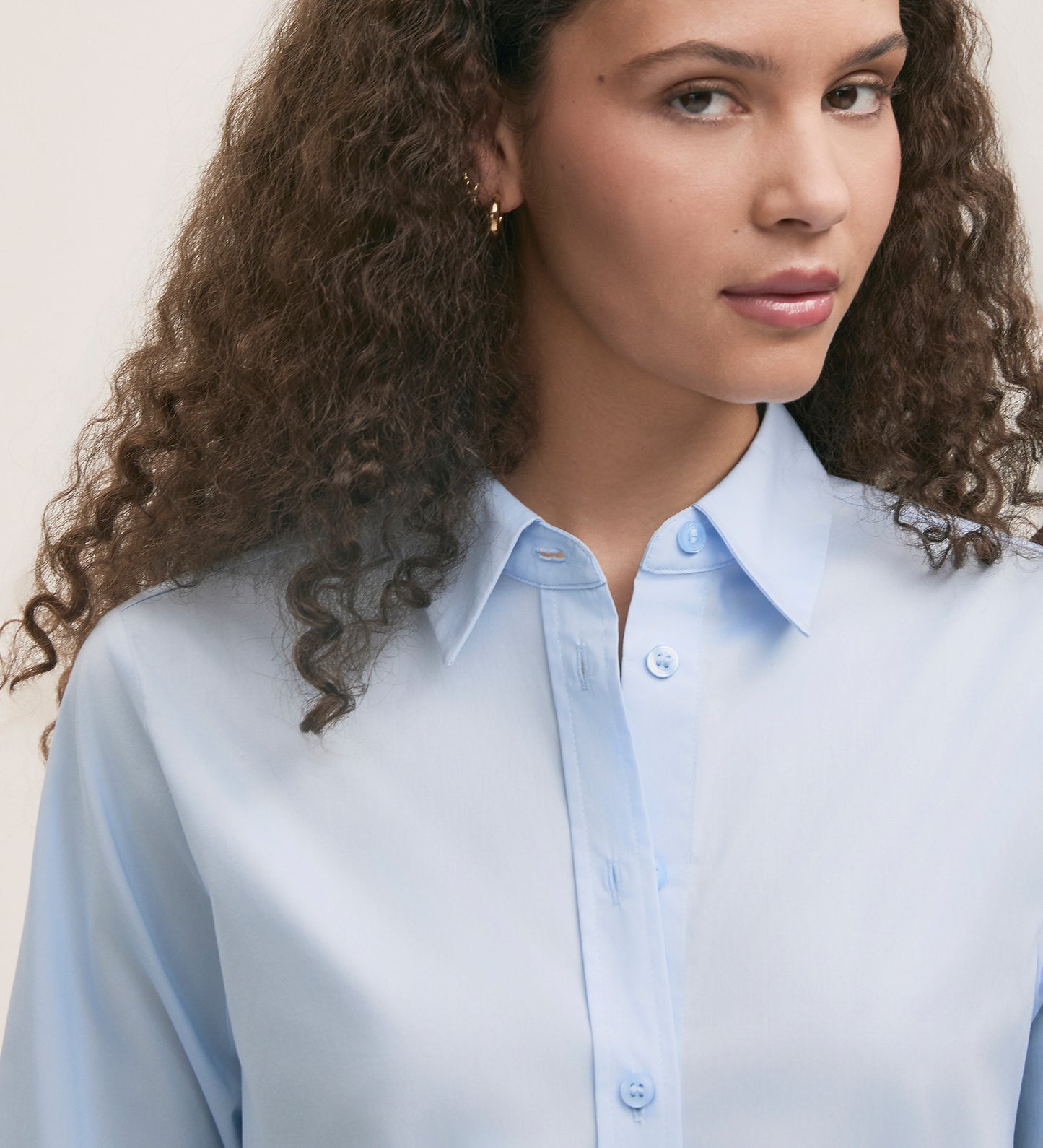 A woman with curly brown hair wears the Dillon Light Blue Cotton Shirt in a regular fit, gazing slightly to the side with a neutral expression against a plain background.