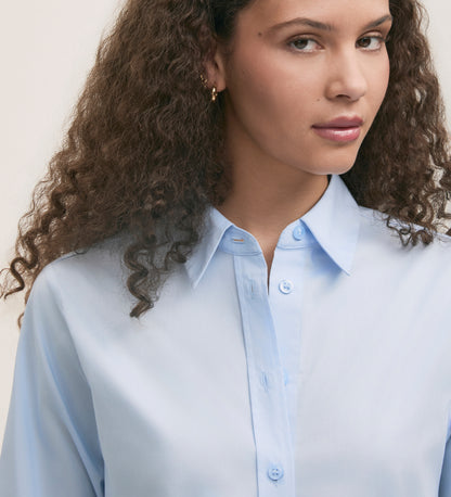 A woman with curly brown hair wears the Dillon Light Blue Cotton Shirt in a regular fit, gazing slightly to the side with a neutral expression against a plain background.