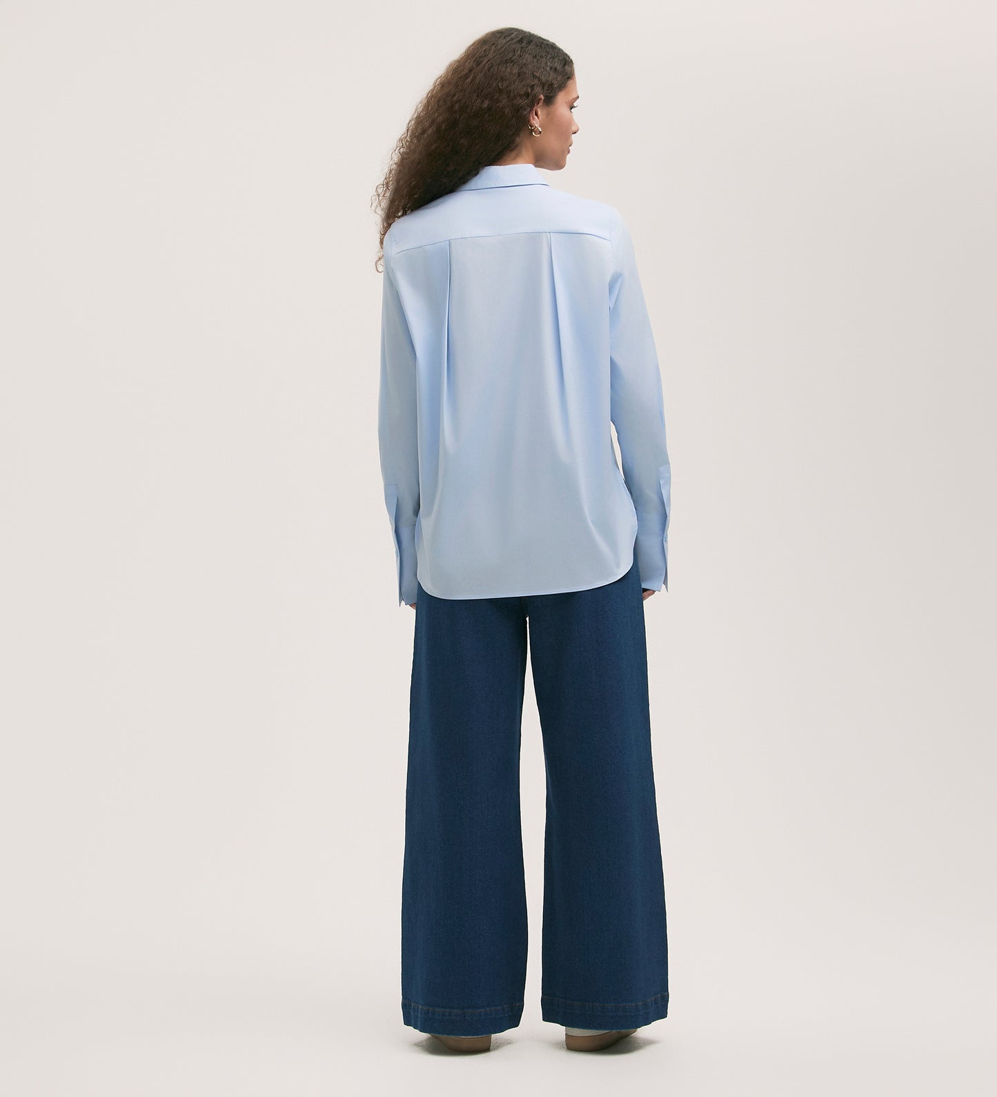 Someone with long curly hair stands facing away on a light background, wearing the Dillon Light Blue Cotton Shirt and regular-fit wide-leg dark blue jeans.