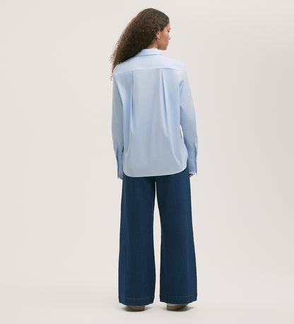 Someone with long curly hair stands facing away on a light background, wearing the Dillon Light Blue Cotton Shirt and regular-fit wide-leg dark blue jeans.