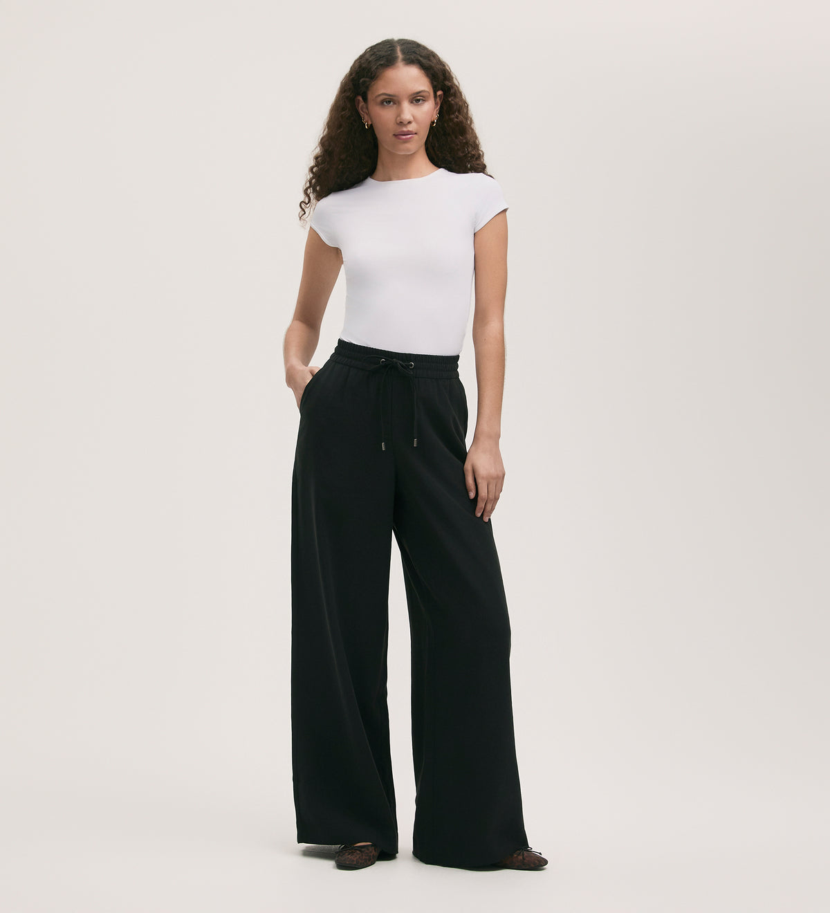 A person with long curly hair wears a fitted short-sleeve white top and Dixie Black Wide Leg Trousers, hands in pockets, standing against a plain light background.