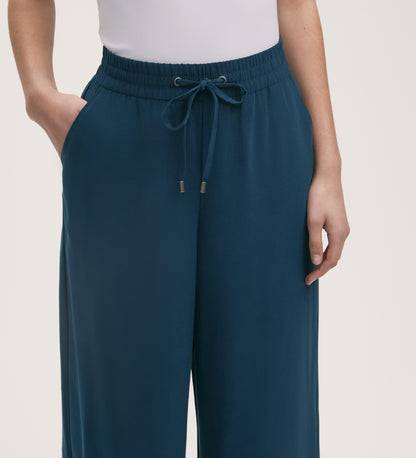 Someone wears Dixie Indigo Wide Leg Trousers featuring a relaxed fit, elasticated waistband, and drawstring, styled with a white sleeveless top. One hand rests in a pocket while the other hangs by their side.