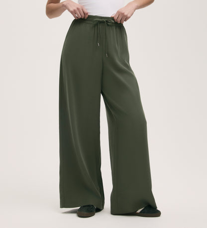 Wearing Dixie Khaki Wide Leg Trousers with a relaxed fit and elasticated waistband, paired with a white top and black shoes, the person stands against a plain light background.