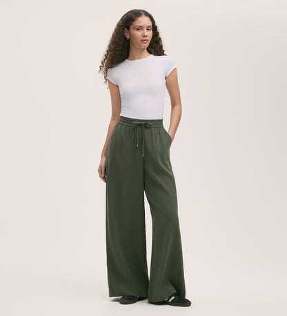 A woman with long curly hair wears a fitted white short-sleeve top, black shoes, and Dixie Khaki Wide Leg Trousers with an elasticated waistband, standing against a plain light background.