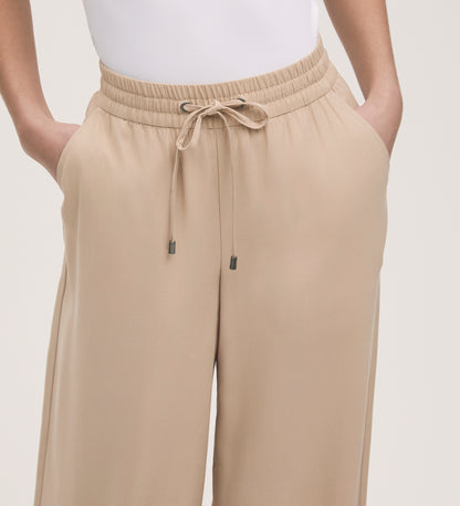 A person wears Dixie Stone Wide Leg Trousers with a relaxed fit, elasticated waistband, and adjustable drawstring, standing hands in pockets while the top of a white shirt peeks out.