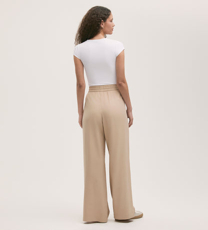 A woman with curly hair stands facing away against a light background, wearing a white short-sleeve fitted top and Dixie Stone Wide Leg Trousers featuring a relaxed fit and elasticated waistband.