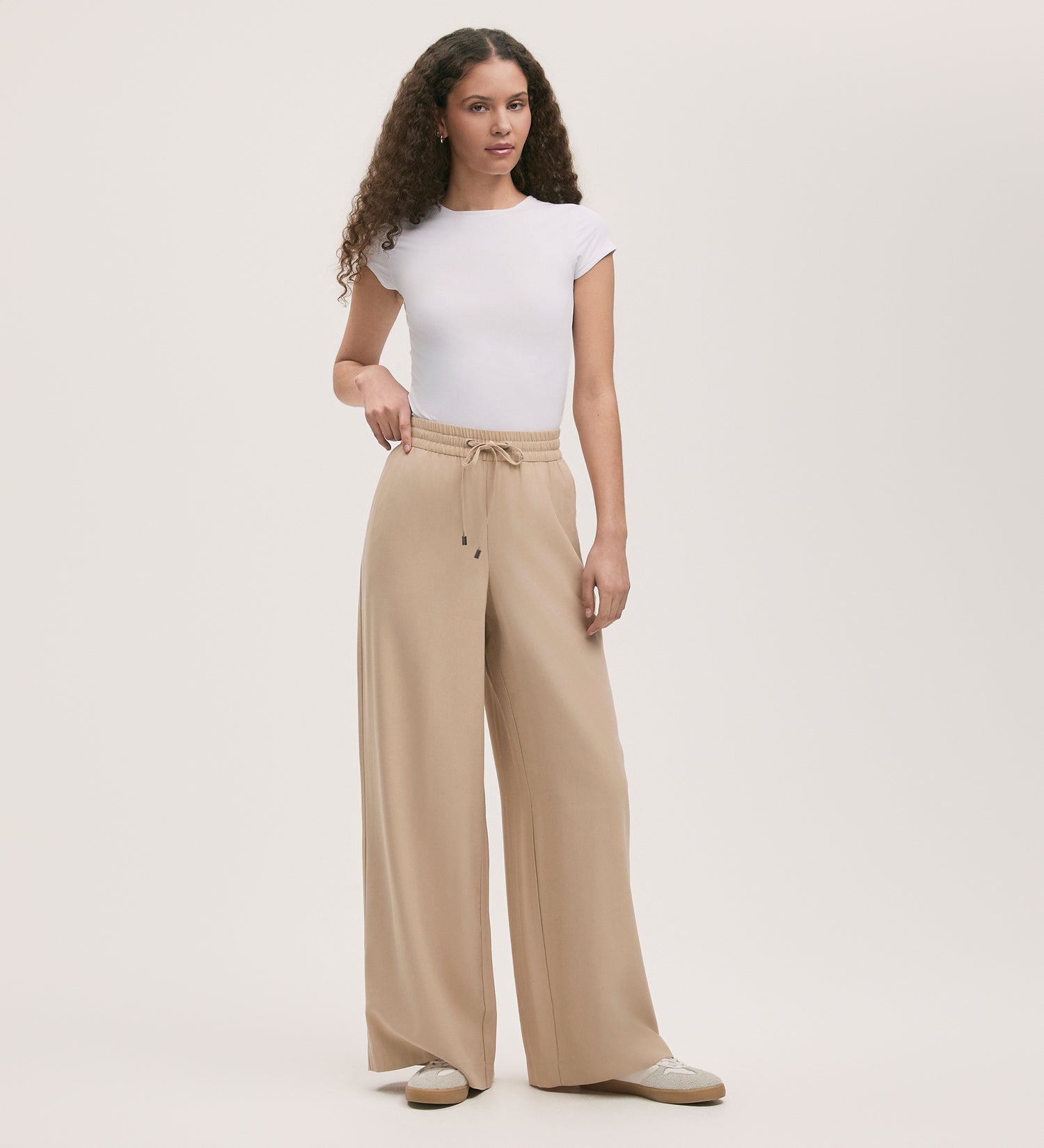 A woman with curly hair poses casually in Dixie Stone Wide Leg Trousers, a fitted white tee, and white sneakers against a plain background. The beige trousers feature a drawstring and elastic waistband for comfort.