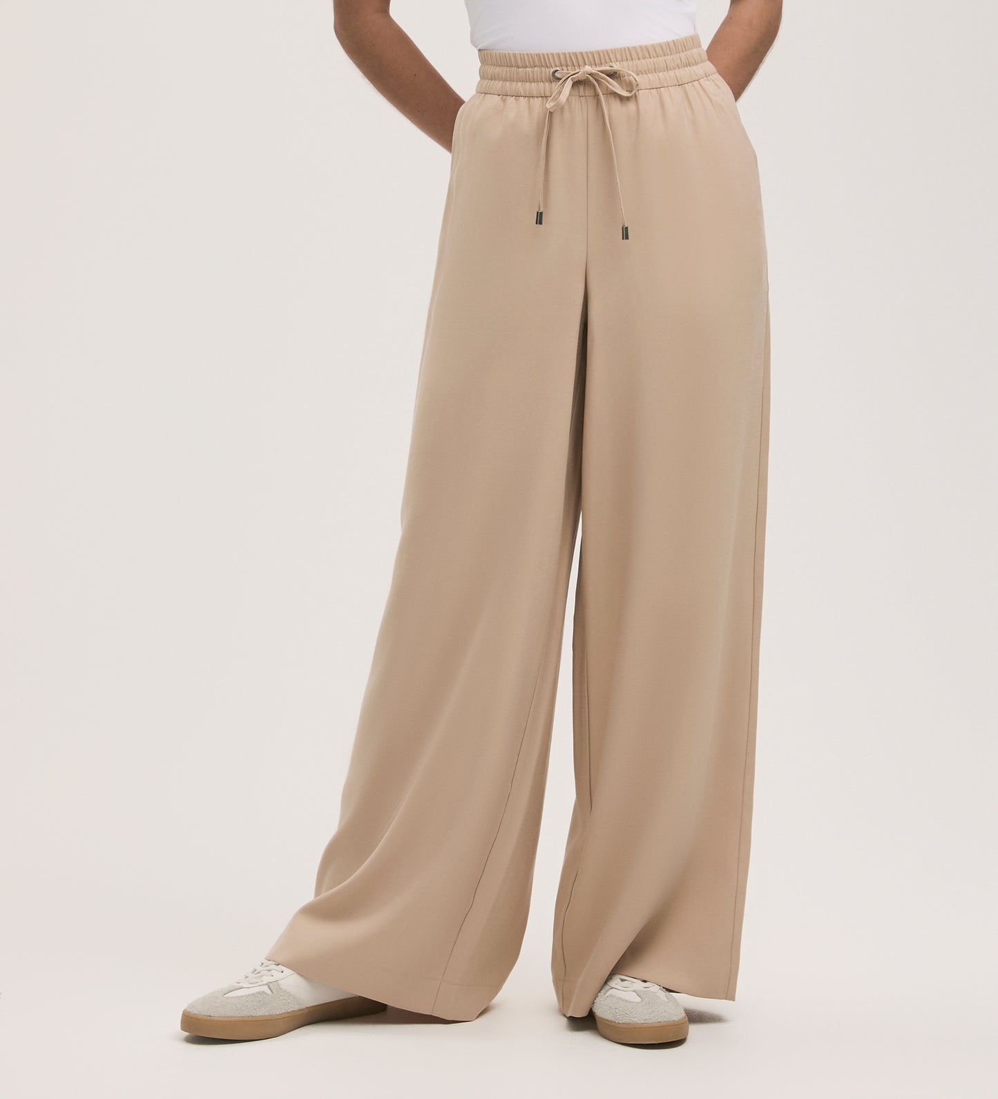 A person stands against a plain backdrop wearing Dixie Stone Wide Leg Trousers with an adjustable drawstring and elasticated waistband, styled with a white top and white sneakers.
