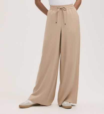 A person stands against a plain backdrop wearing Dixie Stone Wide Leg Trousers with an adjustable drawstring and elasticated waistband, styled with a white top and white sneakers.