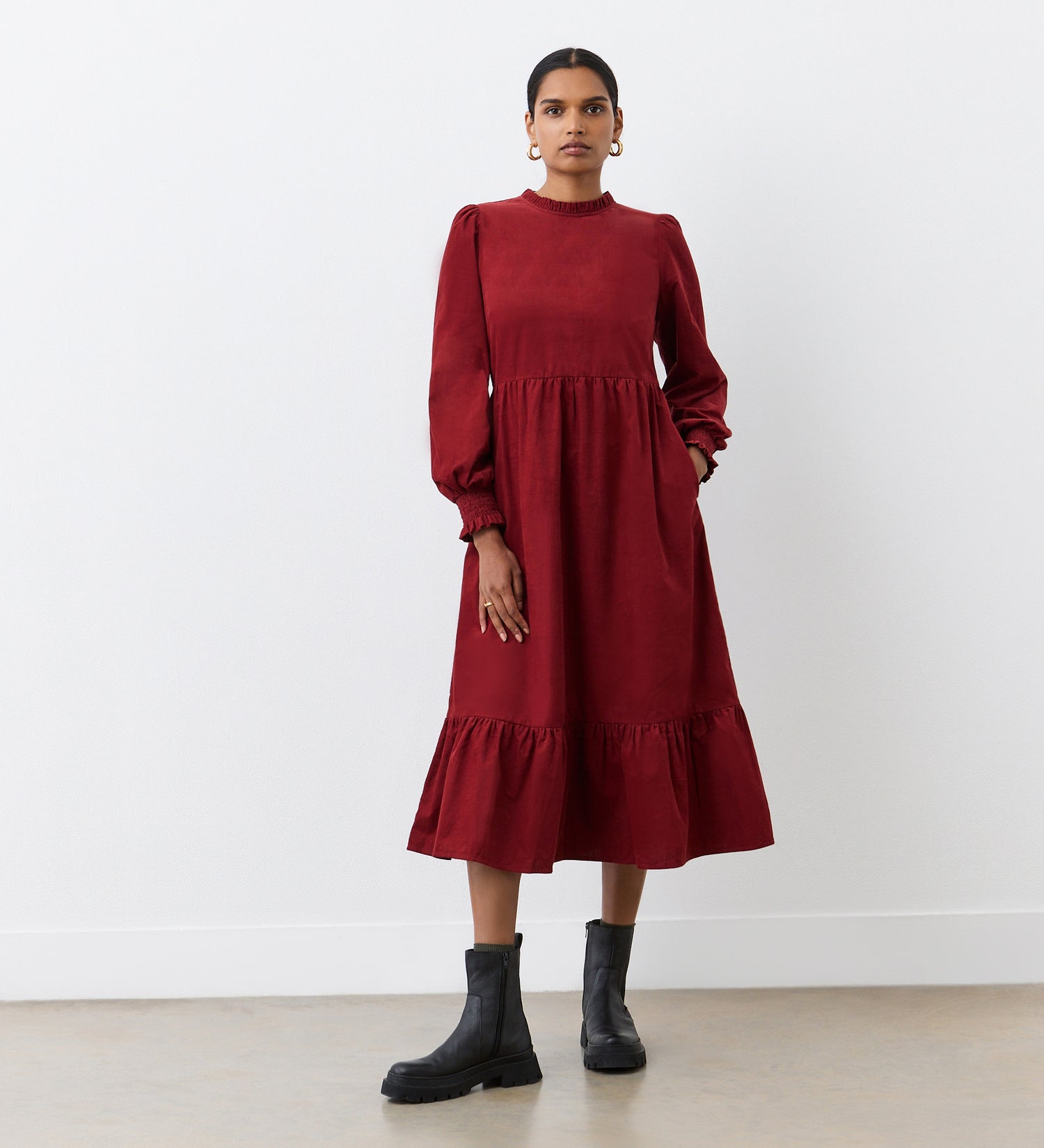 A woman stands against a white wall wearing the Esme Rust Corduroy Midi Dress, featuring long sleeves, ruffled cuffs, and a tiered hemline. She pairs it with black ankle boots, has her hair pulled back, and wears hoop earrings.