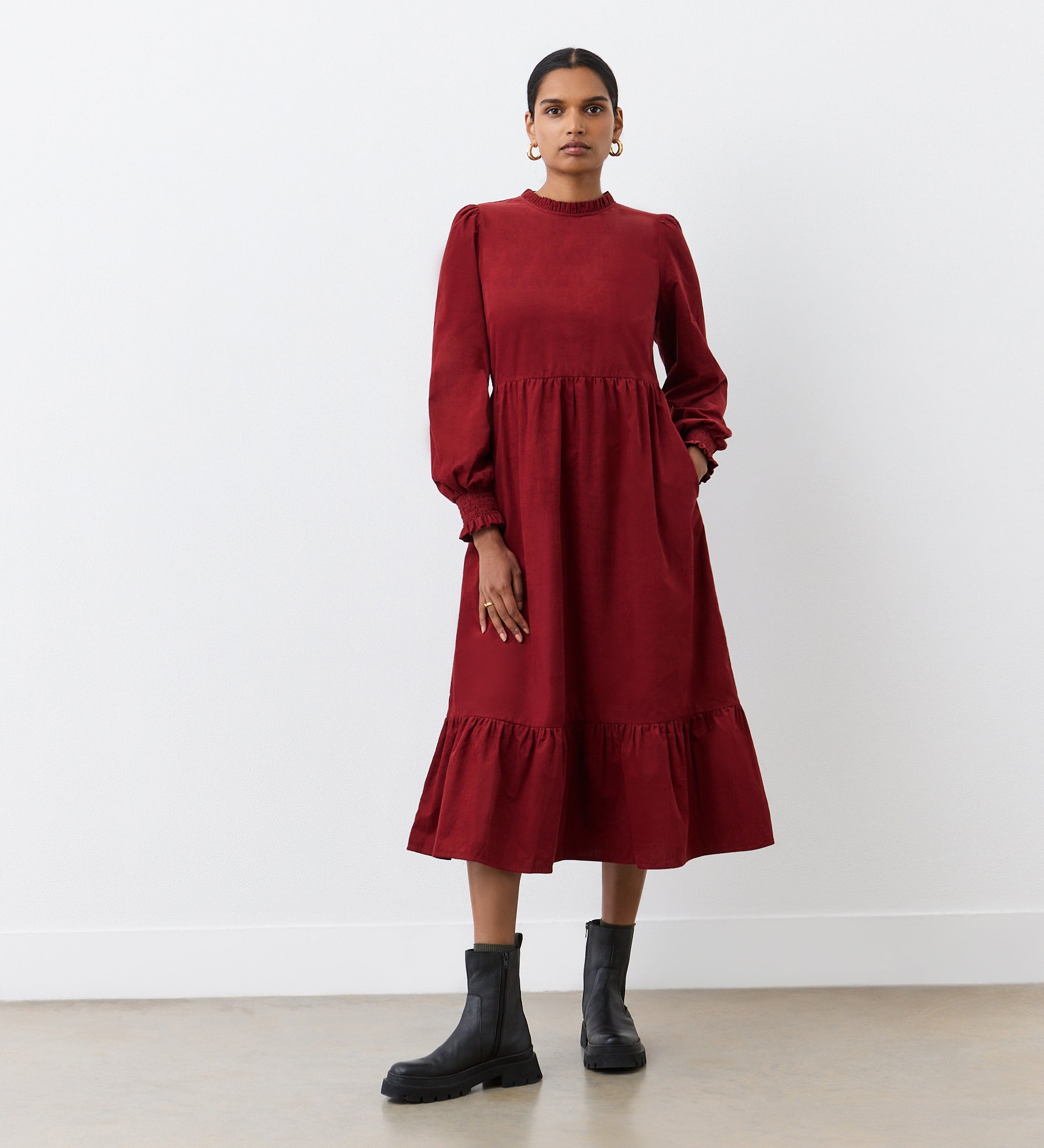 A woman stands against a white wall wearing the Esme Rust Corduroy Midi Dress, featuring long sleeves, ruffled cuffs, and a tiered hemline. She pairs it with black ankle boots, has her hair pulled back, and wears hoop earrings.