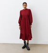 A woman stands against a white wall, wearing the Esme Rust Corduroy Midi Dress with long sleeves and a tiered hemline, paired with black ankle boots. Her hair is pulled back and she accessorizes with hoop earrings.