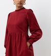 Woman models the Esme Rust Corduroy Midi Dress, featuring long sleeves, a high neck, ruffled cuffs, a tiered hemline, pockets, and rich rust color against a plain white background.