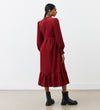 A woman stands facing away in the Esme Rust Corduroy Midi Dress, featuring long sleeves and a tiered hemline. Her hair is tied in a low ponytail, and she pairs the dress with black ankle boots.