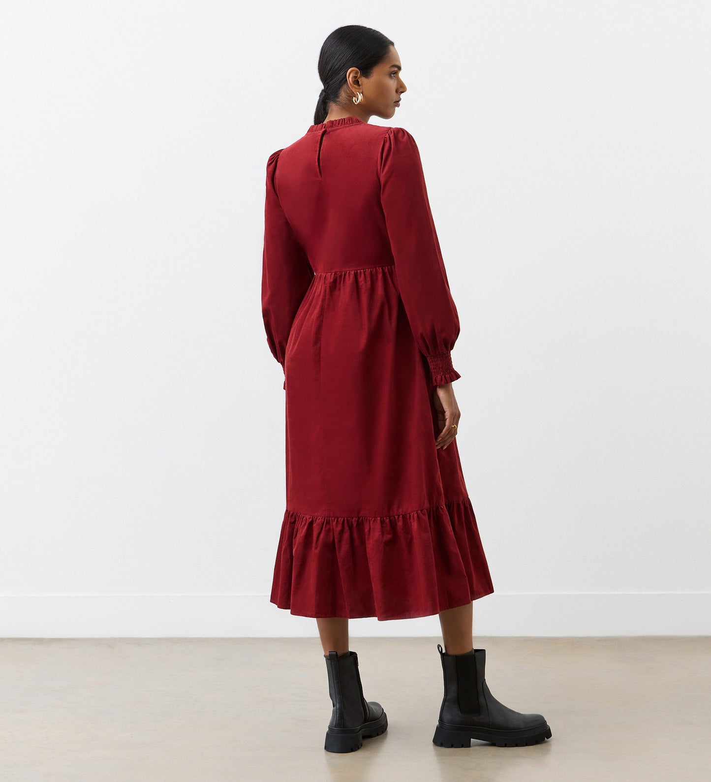 A woman stands facing away in the Esme Rust Corduroy Midi Dress, featuring long sleeves and a tiered hemline. Her hair is tied in a low ponytail, and she pairs the dress with black ankle boots.
