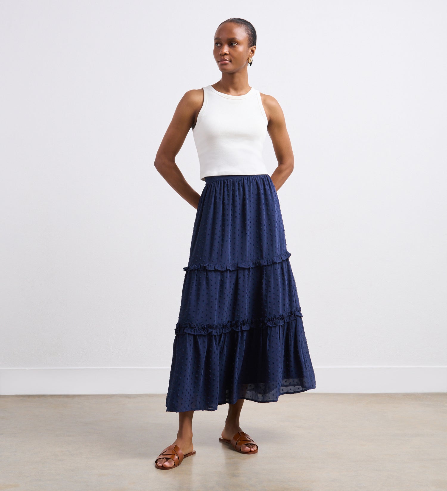 A woman wearing a sleeveless white top and the Everley Navy Midi Skirt stands against a plain white wall, looking to the side with her hands behind her back.