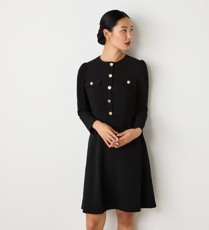 A woman with dark hair in a low bun wears the Evie Black Crepe Short Dress, featuring gold button details and chest pockets, standing against a plain white wall and looking to her left.