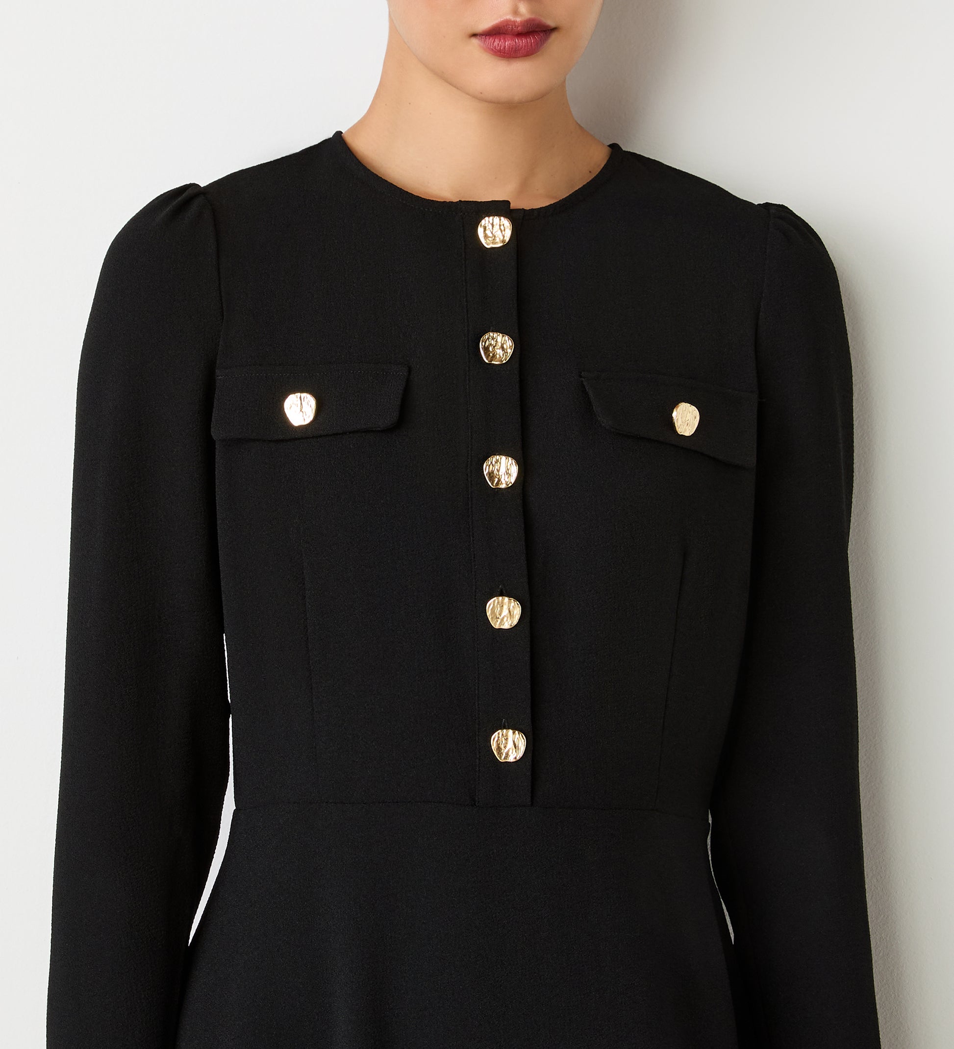 A woman models the Evie Black Crepe Short Dress featuring gold button details down the front and on two chest pockets, standing against a plain white background. Her face is partially shown with neutral makeup and pink lipstick.