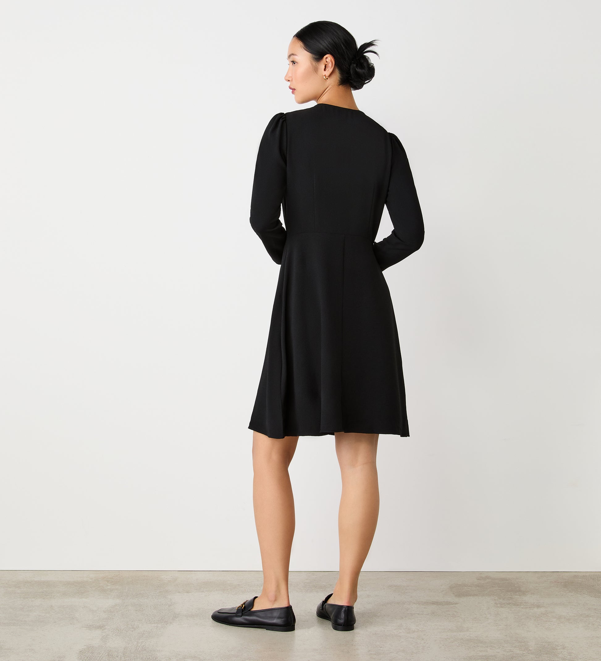 A woman stands with her back to the camera, wearing the Evie Black Crepe Short Dress. Her hair is styled in a neat bun, and she pairs the knee-length dress with black loafers against a plain white wall and concrete floor.