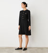 A woman stands indoors against a white wall, wearing the Evie Black Crepe Short Dress with a fitted bodice, long sleeves, and gold button details on the front and pockets. She pairs it with black loafers and has her dark hair tied back.