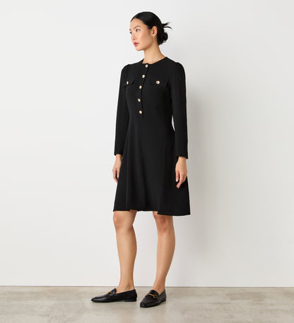 A woman stands indoors against a white wall, wearing the Evie Black Crepe Short Dress with a fitted bodice, long sleeves, and gold button details on the front and pockets. She pairs it with black loafers and has her dark hair tied back.