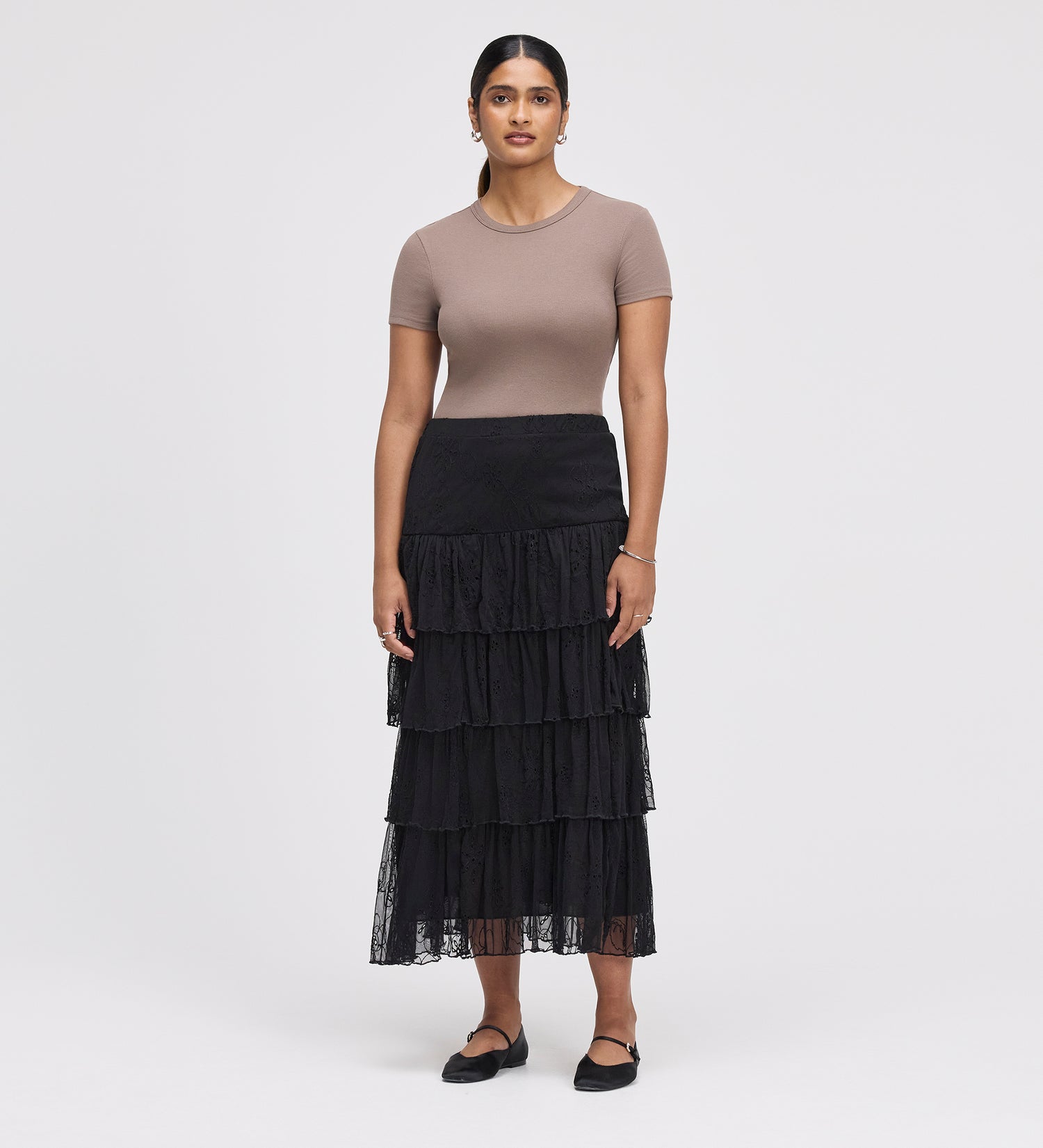 A woman stands against a plain background wearing a fitted taupe short-sleeve top and the Elisa Black Mesh Midi Tiered Skirt with an elastic waistband. She completes the look with black flats, earrings, and a bracelet.