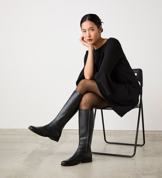 A woman with straight black hair, dressed in a black dress and tights, models the Frankie Black Knee High Leather Boots—an elegant choice for any autumn wardrobe—as she sits on a black folding chair against a plain white background.