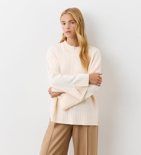 A woman with long blonde hair stands against a white wall, wearing the Frankie Ivory Boxy Jumper with ribbed details and side splits, paired with light brown pants. Her arms are crossed and she has a neutral expression.