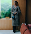 A woman stands indoors wearing Veronica Natural Animal Ruffle Empire Waist Midi Dress holding a dark blue handbag. She is posed beside a window with a wooden wall and a terracotta-colored sofa in a modern interior setting.