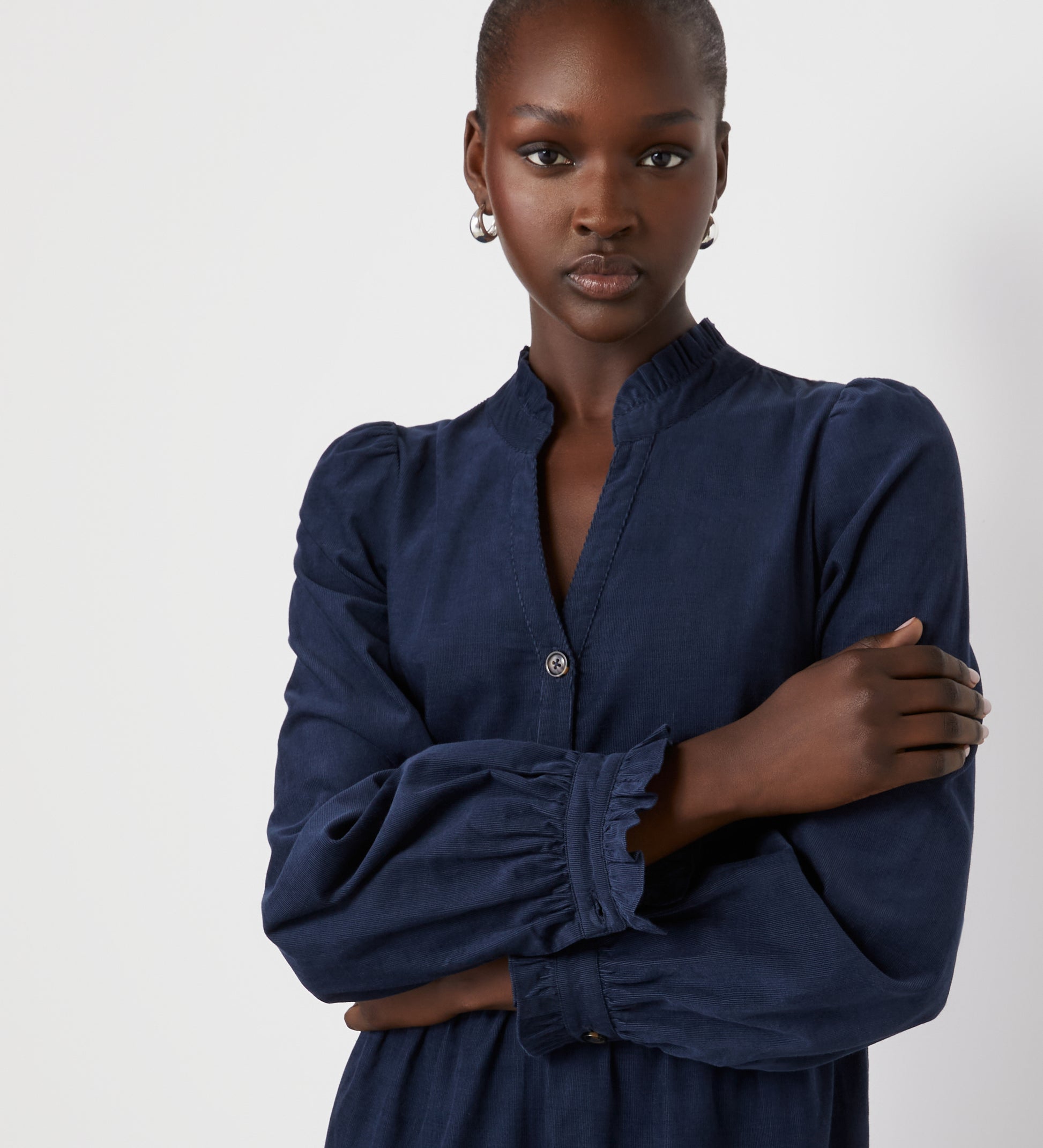 A woman with dark skin and hoop earrings stands against a white background, arms crossed, wearing the Gianna Navy Corduroy Midi Dress with a ruffled collar, front buttons, and a tiered skirt.