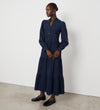 A woman stands against a plain white wall in the Gianna Navy Corduroy Midi Dress, featuring front buttons and a tiered skirt. Her hair is pulled back and she wears black loafers, gazing thoughtfully to the side.