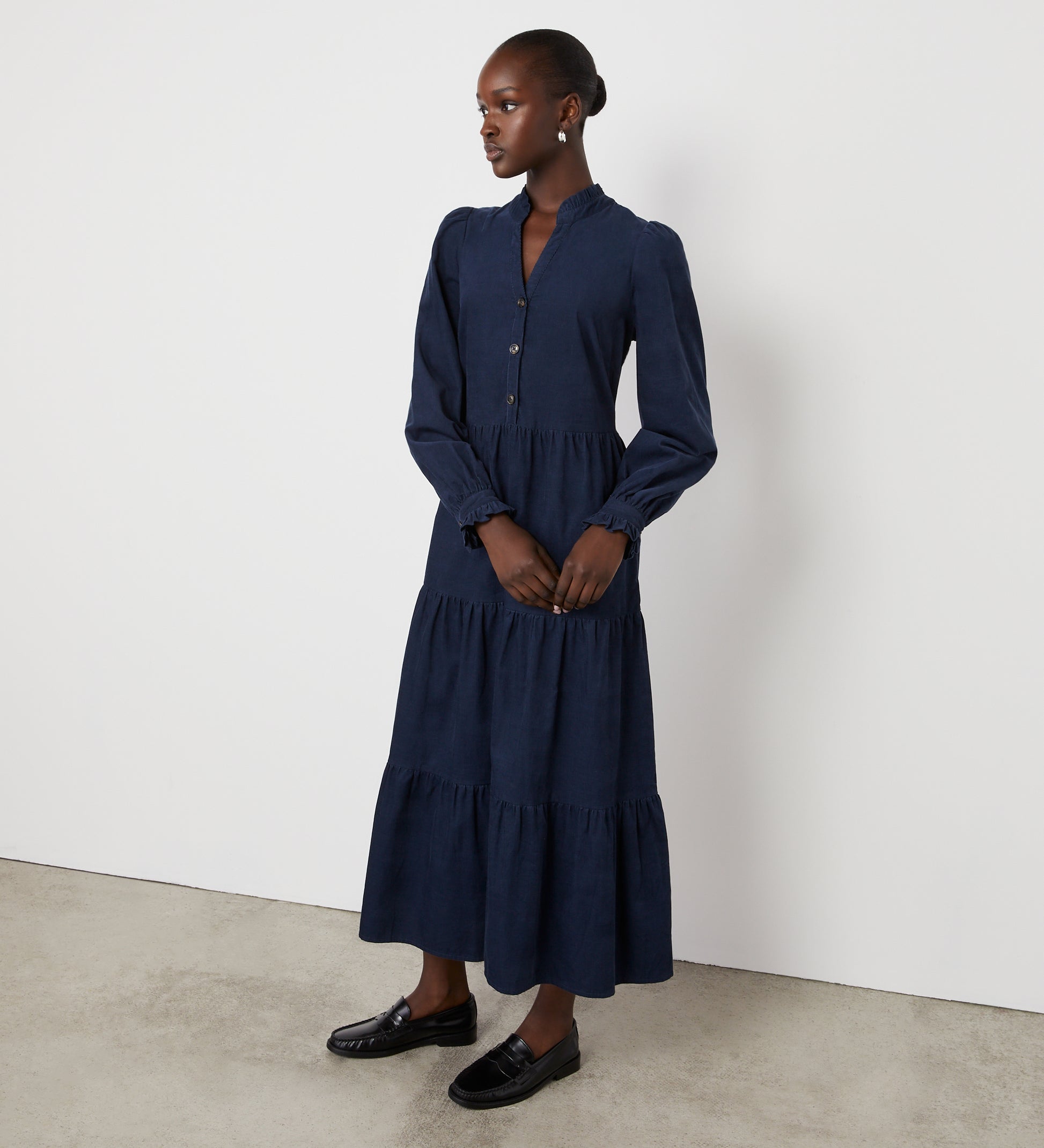 A woman stands against a plain white wall in the Gianna Navy Corduroy Midi Dress, featuring front buttons and a tiered skirt. Her hair is pulled back and she wears black loafers, gazing thoughtfully to the side.