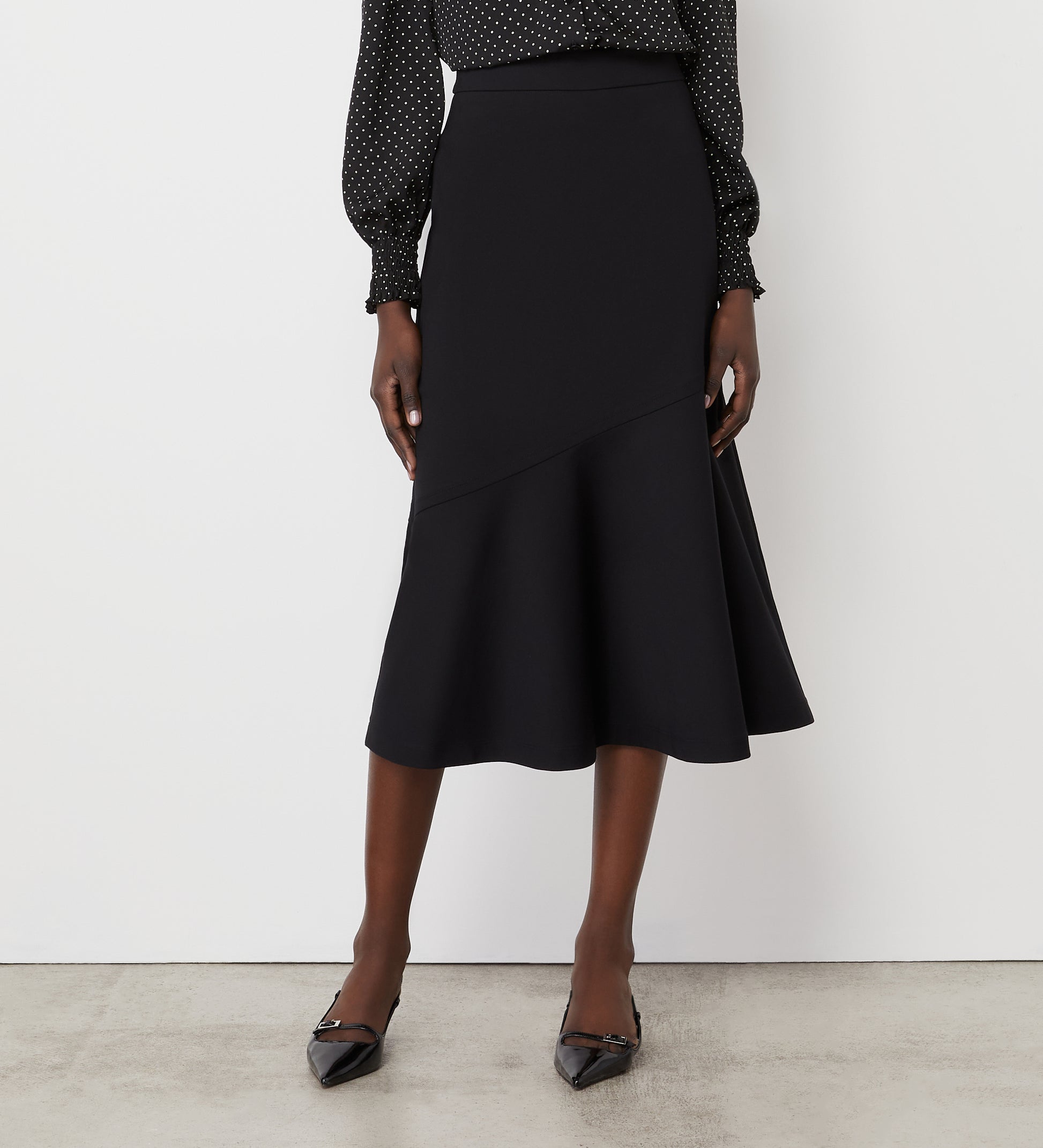 A woman wearing a black, long-sleeve, polka dot blouse and the Grace Black Ponte Jersey Midi Skirt with an asymmetrical hem, paired with black pointed-toe heels, stands against a plain white wall.