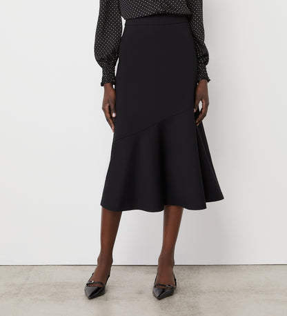 A woman wearing a black, long-sleeve, polka dot blouse and the Grace Black Ponte Jersey Midi Skirt with an asymmetrical hem, paired with black pointed-toe heels, stands against a plain white wall.